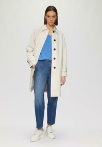 Trench-coat crème avec des boutons noirs, porté sur un haut bleu et un jean bleu. Dispose de manches longues et d'une silhouette droite. Une paire de baskets blanches.