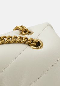Cremefarbene Ledertasche mit einer goldenen Kettenriemen und metallischen Ösen. Auffällige Nähte und subtile Textur auf der Oberfläche.