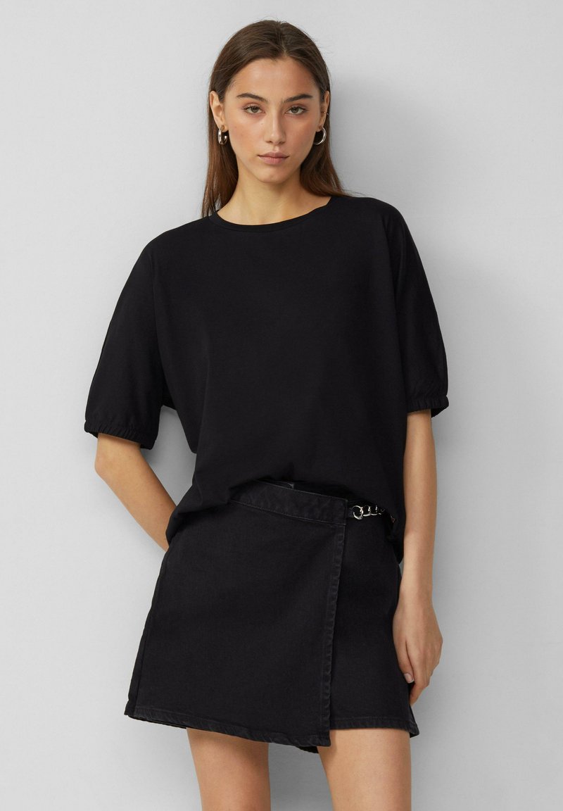 Czarna bluzka krótko rękawowa z okrągłym dekoltem i detalami przy mankietach, połączona z czarną dżinsową spódnicą o kroju kopertowym z paskiem i metalowymi akcentami.