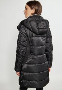Geacă puffer neagră cu glugă, având un design matlasat, textură lucioasă și panouri din plasă pe laterale. Lungime medie, cu talie strânsă.