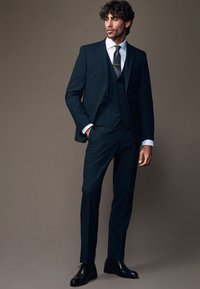 Marineblauer Dreiteiler mit taillierter Passform, einreihiger Jacke, Reverskragen und passender Hose. Schwarze Abendkle Schuhe runden den Look ab.