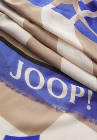 Écharpe multicolore avec un motif géométrique comportant des sections bleu, beige et blanc. Logo de la marque "JOOP!" en blanc audacieux sur une bande bleue. Tissu texturé avec des bords effilochés.