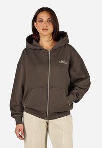 Reißverschluss-Hoodie aus dunkelbraunem Baumwoll-Mix, mit einer Fronttasche und gesticktem "Cozy Club"-Logo in Weiß auf der Brust.