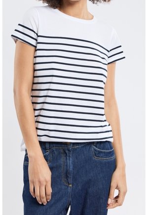 Frau trägt ein weißes Kurzarmshirt mit schwarzen horizontalen Streifen und blaue Jeans, steht vor einem einfarbigen Hintergrund.