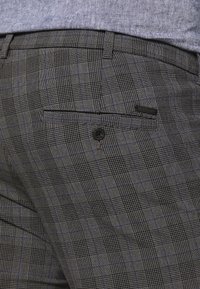 Close-up van grijze geruite broeken met een zwarte knoop achterzak, gedragen met een blauwe shirt.