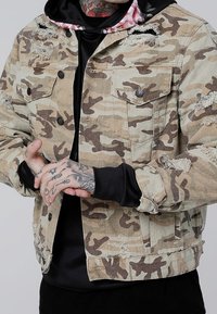 Kamuflaasi denim takki, jossa on kulutettuja yksityiskohtia, nappilukkoja ja rintataskuja. Päällä musta huppari, jossa on kukkakoristelu.