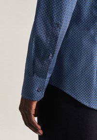 Mörkhyad hand vilande vid sidan iförd en blå långärmad skjorta med geometriska mönster och svarta byxor mot en enfärgad bakgrund.