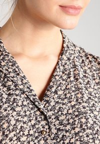 Detailní záběr na krk a bradu osoby, která má na sobě košili s květinovým vzorem s límečkem a knoflíkem. Měkké, přirozené osvětlení.