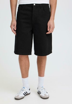 Man wearing black knee-length shorts, white socks, and white sneakers with black stripes, standing against a plain background.