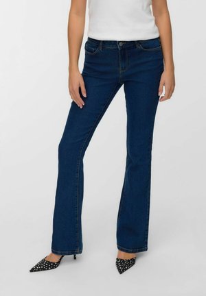 Femme portant un jean bootcut bleu foncé, un haut sans manches blanc et des escarpins noirs à pois blancs, se tenant contre un fond uni.