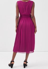 Robe fuchsia plissée avec une ceinture texturée, ourlet longueur genou et design sans manches. Portée avec des sandales à talons carrés noires.
