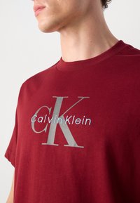 Burgundijasti pamukasti majica s velikim sivoj "CK" logotipom i manjim "Calvin Klein" tekstom u svijetloplavoj boji. Kratki rukavi i okrugli izrez.