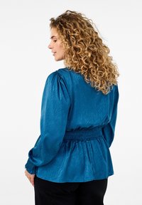 Blouse turquoise en tissu texturé, taille smockée et manches bouffantes. La vue arrière présente un doux éclat et un motif ondulé subtil.