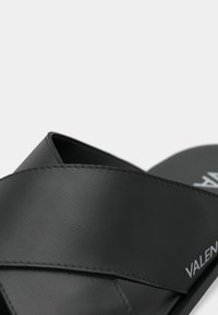 Svart slidesandal med kryssremdesign, laget av glatt syntetisk materiale. Merkenavnet "VALENTINO" vises på fotsengen.