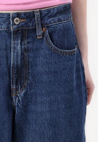 Jeans de mezclilla azules con cintura alta, costura visible y un bolsillo delantero. Presentan un cierre de botón y un acabado de tela texturizada.