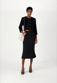 Nais, kes kannab musta lühikest nööpidega jope, musta midi seelikut, musti kontsakingi ja hoiab käes valget õlakotilt, seistes lihtsa valge tausta ees.