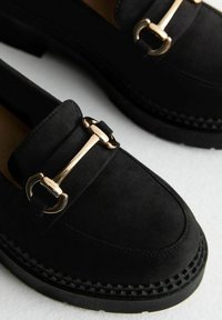 Los mocasines de ante negro presentan un acento de hardware en tono dorado y una suela texturizada con un diseño trenzado. Superficie lisa con forma de punta redondeada.