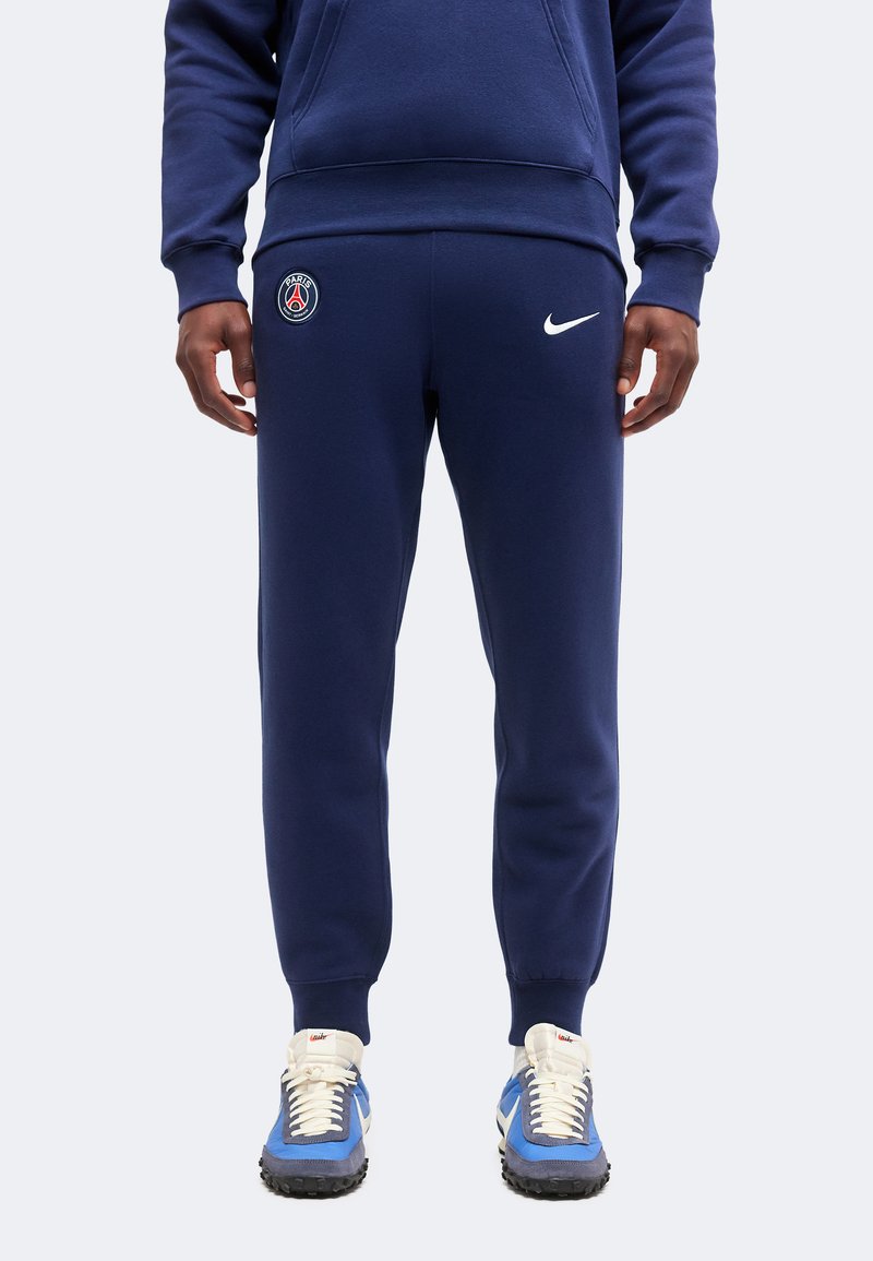 Άνδρας που φοράει σκούρο μπλε φόρμα Paris Saint-Germain με λογότυπο Nike, σε συνδυασμό με μπλε και λευκά αθλητικά παπούτσια, στέκεται μπροστά σε μονόχρωμο φόντο.