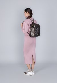 Mochila marrón acolchada llevada en la espalda, combinada con un vestido de manga larga rosa pálido con una abertura lateral y zapatillas de color claro.