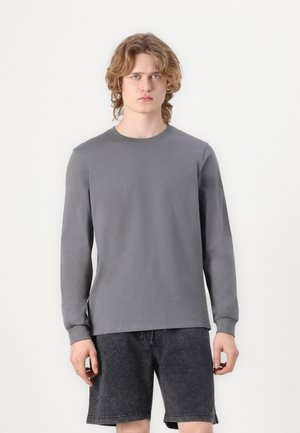 T-shirt gris à manches longues en tissu lisse, avec un col rond et des manches ajustées, associé à un short texturé sombre.