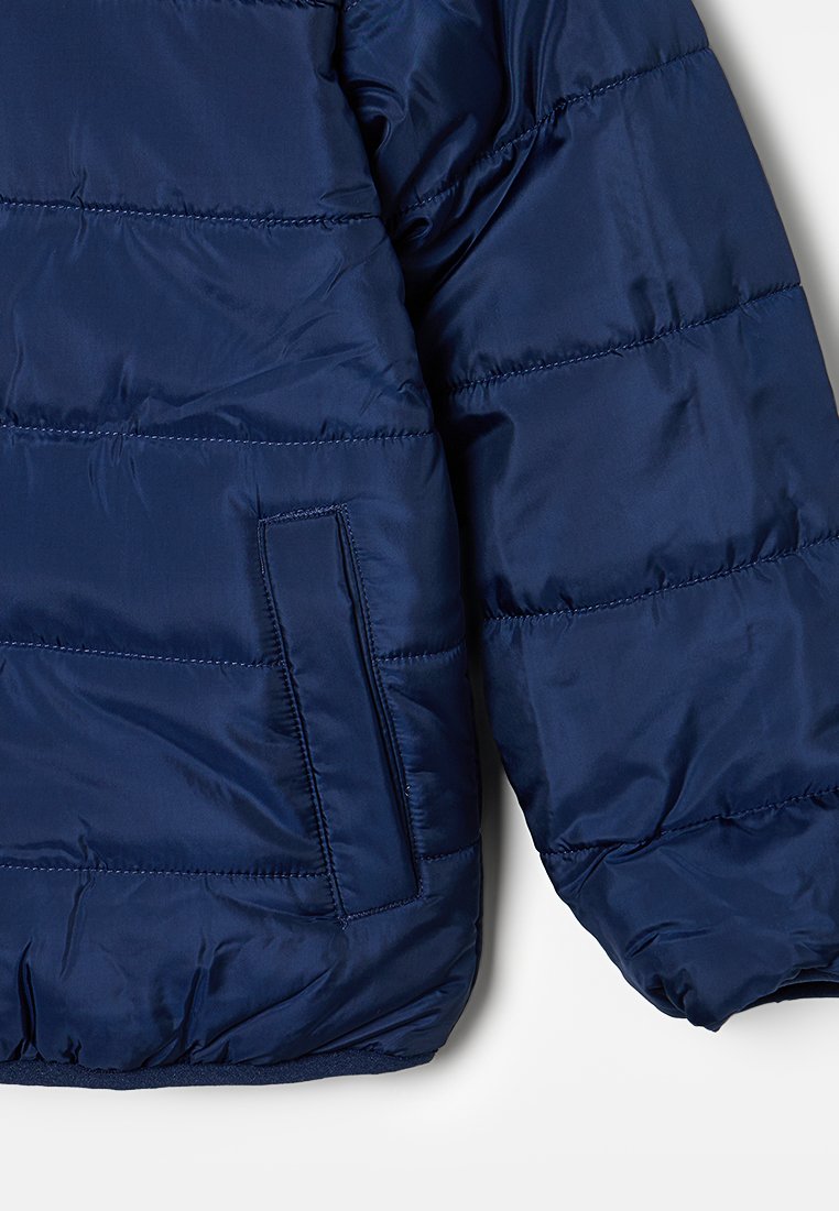 Veste rembourrée bleu marine avec texture matelassée, dotée d'une poche latérale et de poignets élastiques. Fabriquée en tissu léger et brillant.