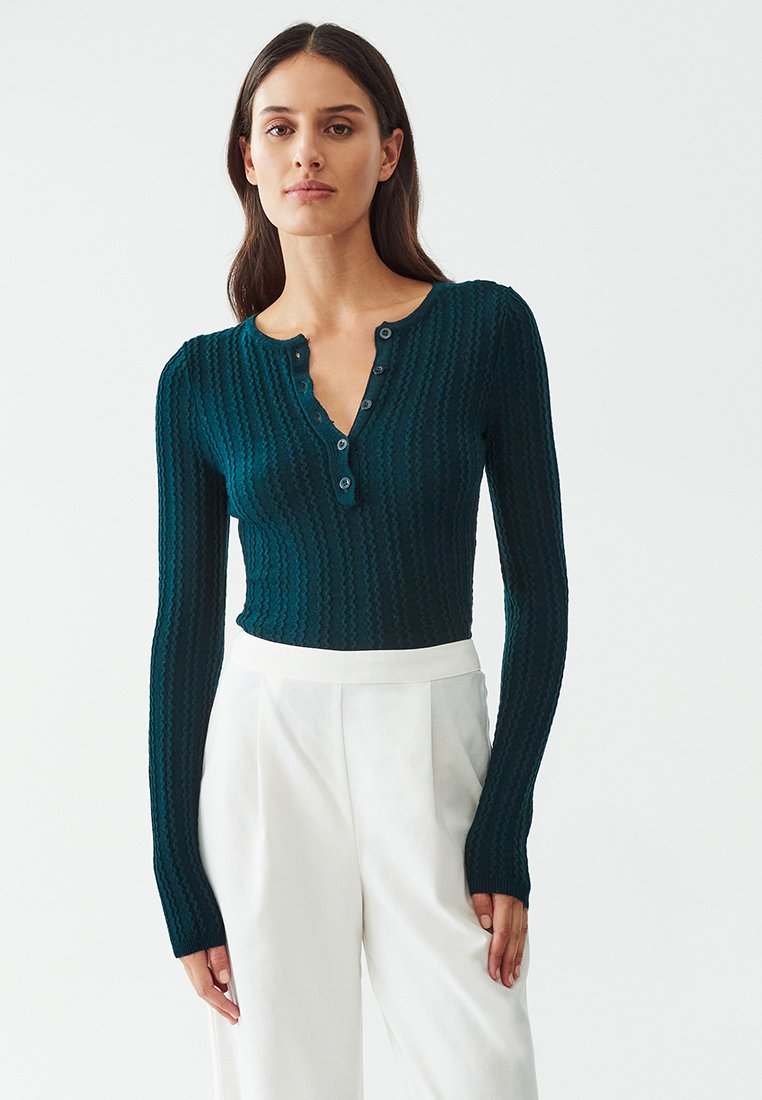 Teal ribbed long-sleeve top with a henley neckline, featuring three buttons and a fitted design, paired with high-waisted white trousers.