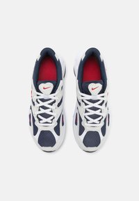 Nike sportbačiai su balta tinkline viršutine dalimi, tamsiai mėlynomis detalėmis, raudonais akcentais ant liežuvėlio ir tekstūruotu padu. Yra baltos raištelės ir logotipas.