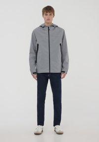 Giacca impermeabile grigio chiaro con cappuccio, zip frontale e tasche laterali con zip, abbinata a jeans blu scuro e sneakers bianche.