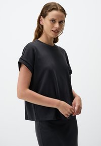 Czarny krótki top z luźnym krojem i podwiniętymi rękawami, wykonany z gładkiego materiału. Model trzyma dolny brzeg obiema rękami.