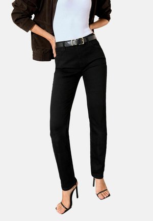 Kvinde iført sorte højtaljede jeans med sort bælte, hvid top, brun jakke og sorte højhælede sandaler med stropper, stående med hænderne på hofterne.