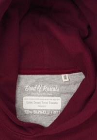 Burgundy bomullshoodie med grå etikett på insidan. Innehåller varumärkesnamn, skötselråd och detaljer om ekologisk textilstandard.