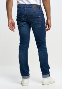 Pantalones vaqueros de mezclilla azul oscuro con un ajuste slim, que presentan bolsillos traseros y un parche de logo de cuero. Con puños enrollados con un ribete de mezclilla más clara.