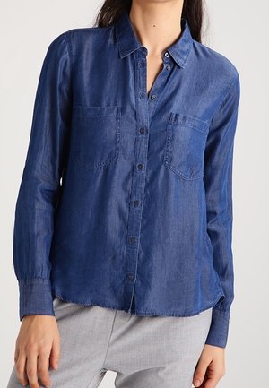 Mujer con una camisa de mezclilla azul de manga larga abotonada, con dos bolsillos en el pecho, y pantalones gris claro.