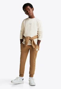 Felpa crema con patch del logo, pantaloni della tuta marroni con tasche e sneakers bianche. Un maglione marrone legato in vita aggiunge contrasto.