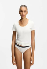 Kvinde med fletninger iført en hvid kortærmet top med rund hals og hvide underbukser med en sort linning mærket "HUGO."