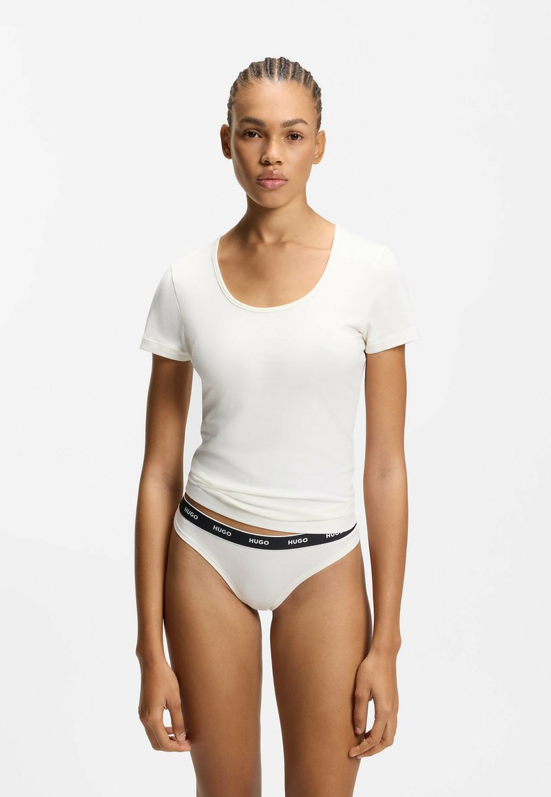 Kvinde med fletninger iført en hvid kortærmet top med rund hals og hvide underbukser med en sort linning mærket "HUGO."