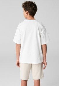 Vit bomull t-shirt med kort ärm, avslappnad passform, rund halsringning, bärs med ljusbeige shorts, sedd bakifrån.