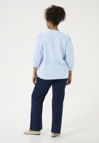 Blouse bleu clair avec des pois texturés, manches ballons et col rond. Associée à un pantalon en denim foncé à jambe droite et des ballerines beiges.