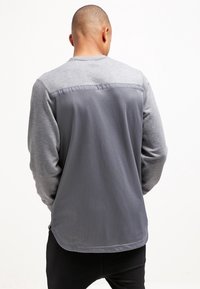 Grå atletisk sweatshirt med långa ärmar. Har en texturerad meshbak, slät framdel och kontrasterande tyg vid axlarna.