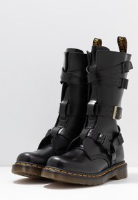 Bottes hautes en cuir noir avec sangles et boucles, embout renforcé et couture jaune sur la semelle en caoutchouc. Finition mate ; conception robuste.