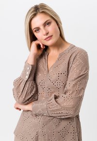 Beige blouse met een lichtgewicht, geborduurd ontwerp met uitsnijdingen, knoopmanchetten en een V-hals. Getextureerde, ademende stof.