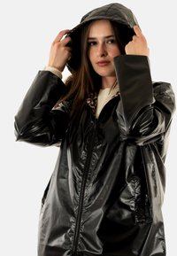 Chaqueta negra impermeable con un acabado brillante, que cuenta con capucha y forro de leopardo. Tiene una cremallera central y bolsillos laterales.