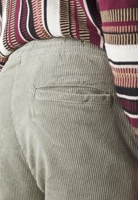 Gros plan sur la poche arrière d'un pantalon en velours côtelé gris porté avec un pull en maille rayé multicolore en bordeaux, blanc, noir et beige.