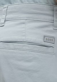 Poche arrière d’un pantalon gris clair avec une fente horizontale et une petite étiquette rectangulaire en tissu "BOSS" cousue au-dessus de la poche sur le côté droit.