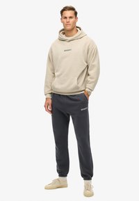 Sudadera con capucha beige con un bolsillo frontal, combinada con pantalones de chándal gris oscuro con una cinturilla y puños elásticos. Ambos artículos son casuales.