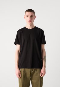 Black cotton t-shirt with a crew neckline and short sleeves, paired with olive green trousers, worn by a person against a neutral background.