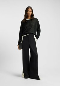 Black sheer long-sleeve shirt with wide neckline, paired with wide-leg black trousers featuring white side panels. Minimalist black handbag.
