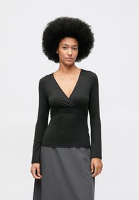 Vrouw met een grote afro-kapsel, gekleed in een zwarte lange mouw diepe V-hals top met kantdetails en een grijze rok, staande tegen een effen achtergrond.