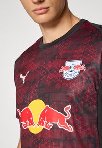 Maillot de football rouge et noir avec un motif géométrique, un logo rond jaune de Red Bull et un logo blanc de Puma sur le tissu.