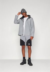 Ung mann iført en grå jakke, matchende lue, svarte og grå shorts, svarte sokker og svarte tursko, står og smiler.